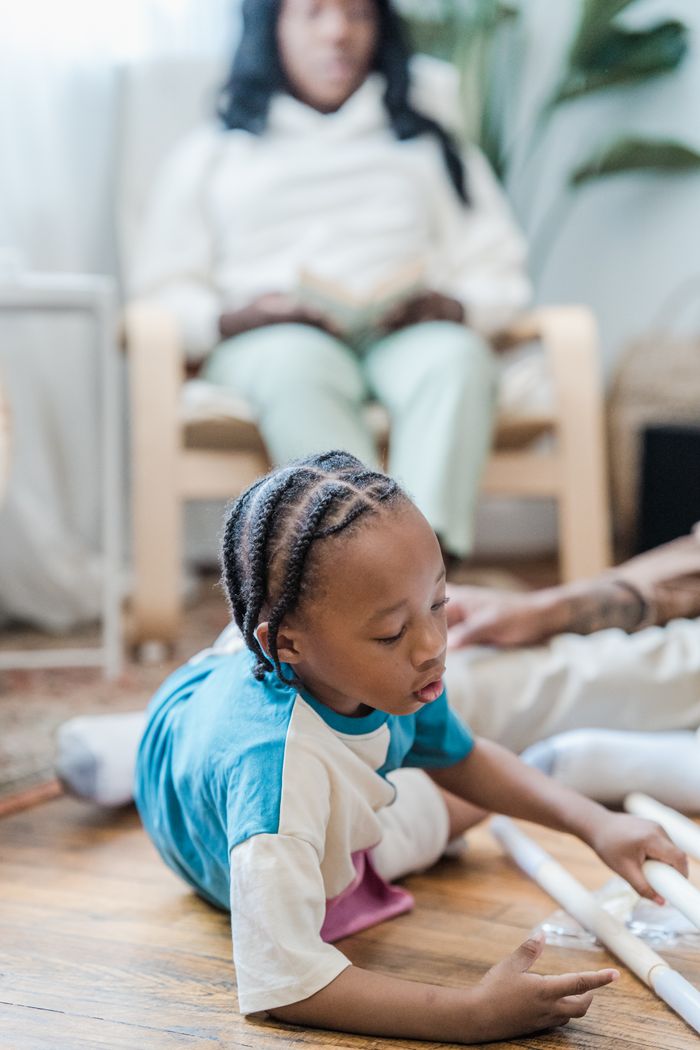 A young boy with cornrows plays on a wooden floor, with a mother figure watching in the background.